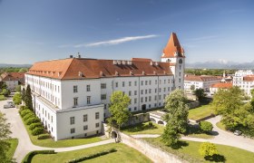 Blick auf eine historische Milit&auml;rakademie mit rotem Dach und Turm, umgeben von gr&uuml;nen B&auml;umen und Rasenfl&auml;chen, im Hintergrund Berge.