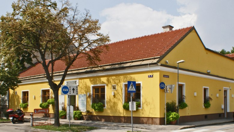 Gelbes Gebäude mit rotem Dach, beschildert als Gasthaus Weidinger, an einer Straßenecke mit Zebrastreifen und Verkehrsschildern.