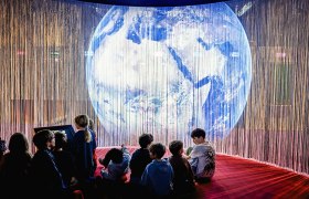 Kinder sitzen vor leuchtender Weltkugel-Projektion in einer Ausstellung.