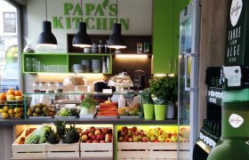 Innenansicht eines modernen Lokals mit frischem Obst und Gemüse, grüner Dekoration und dem Schriftzug 'Papa's Kitchen'.