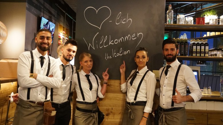 Five people in restaurant clothes stand in front of a board with the inscription ‘Welcome to the Italian’.