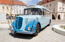 Ein blauer Oldtimer-Bus steht auf einem gepflasterten Platz vor historischen Geb&auml;uden.