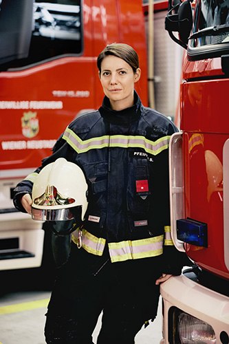 Feuerwehrfrau mit Helm steht im Depot neben mehreren roten Feuerwehrautos.