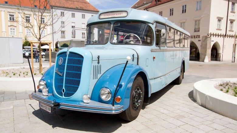 Ein blauer Oldtimer-Bus steht auf einem gepflasterten Platz vor historischen Gebäuden.