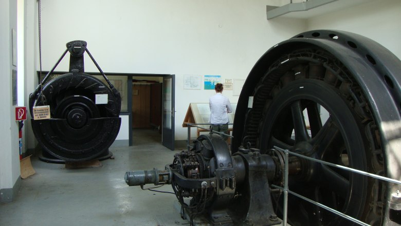 Innenansicht eines Schaukraftwerks mit großen Turbinen und einer Person, die eine Informationstafel liest.