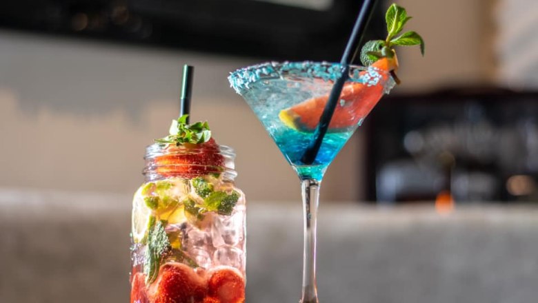 Zwei Cocktails, einer in einem Einmachglas mit Erdbeeren und Minze, der andere in einem Martiniglas mit blauer Flüssigkeit und Zitrusfrucht.
