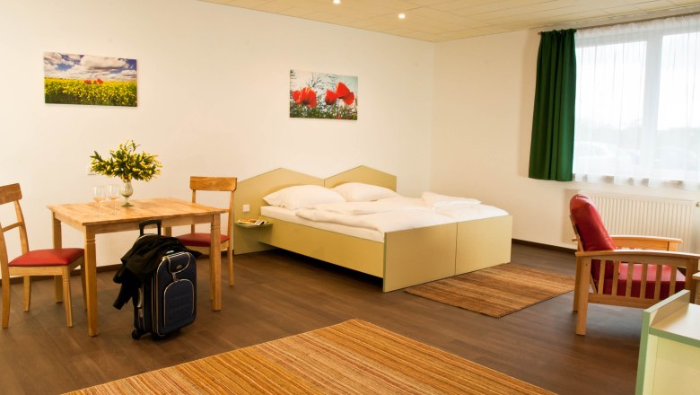 Ein modernes Studio mit Doppelbett, Tisch und Stühlen, rotem Sessel und Koffer. Helle Wände, grüne Vorhänge und Blumenbilder an der Wand.