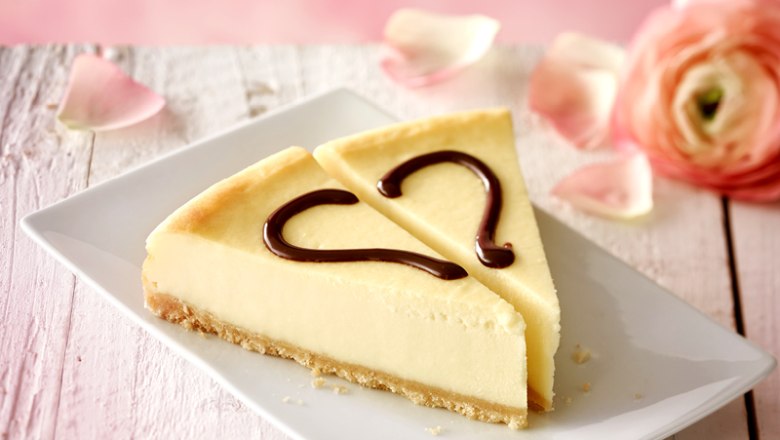 Zwei Stücke Käsekuchen mit Schokoladenherzen auf einem weißen Teller, dekoriert mit Rosenblättern.