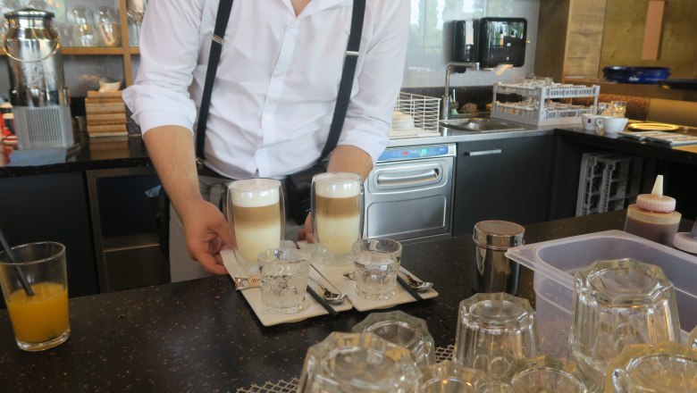 Barista serving two latte macchiatos with glasses of water on a tray in a café.