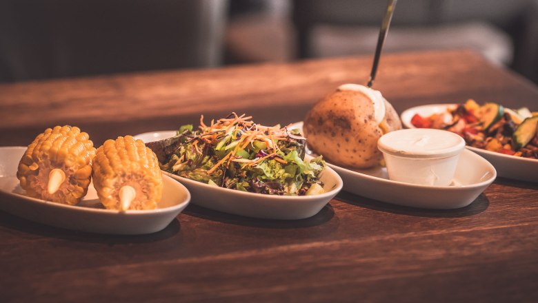 Various side dishes on a wooden table, including corn on the cob, salad, baked potato and dip.